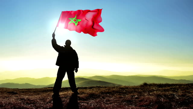 Ganador-de-hombre-exitoso-silueta-ondeando-la-bandera-de-Marruecos-en-la-parte-superior-del-pico-de-la-montaña,-fondo-Cinemagraph-lazo