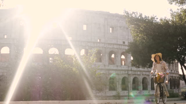 Beautiful-young-woman-in-colorful-fashion-riding-bike-in-front-of-colosseum-in-Rome-at-sunset-with-trees-happy-attractive-girl-tourist-with-straw-hat-in-colle-oppio-front-view-steadycam-dolly