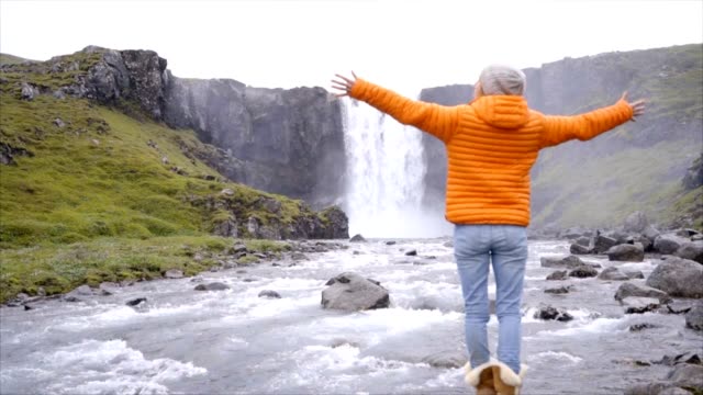 Junge-Frau-vor-dem-herrlichen-Wasserfall-in-Island-ausgestreckten-Armen.-Die-Leute-reisen-Exploration-Konzept-Zeitlupe