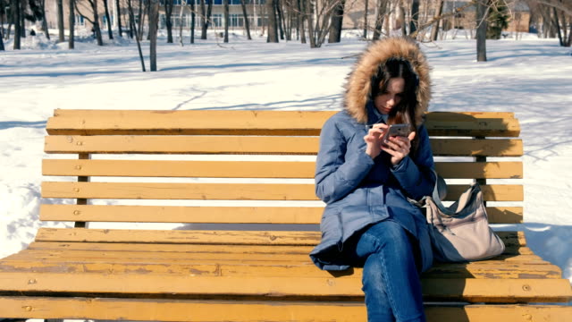Woman-types-a-message-on-her-phone-sitting-on-the-bench-in-winter-city-park-in-sunny-day.