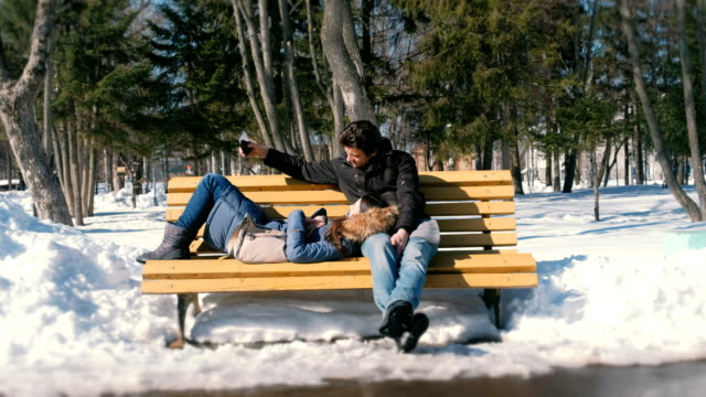 Hombre-y-una-mujer-estar-juntos-en-un-banco-en-la-ciudad-de-winter-Park.-Día-soleado-de-invierno.-Hombre-hace-selfie-en-teléfono.