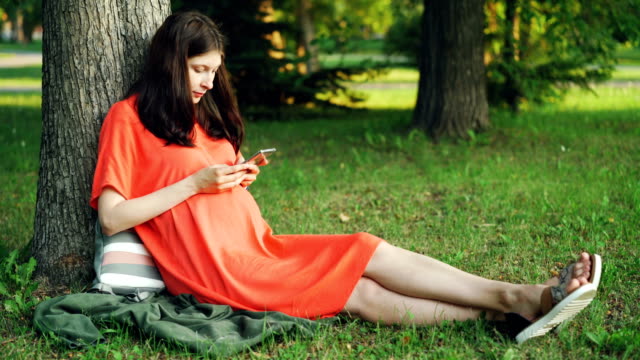 Attraktive-schwangere-Frau-in-Freizeitkleidung-nutzt-Smartphone-sitzen-auf-Rasen-unter-Baum-im-Park.-Schwangerschaft,-Menschen,-moderne-Technology-und-Entspannungs-Konzept.