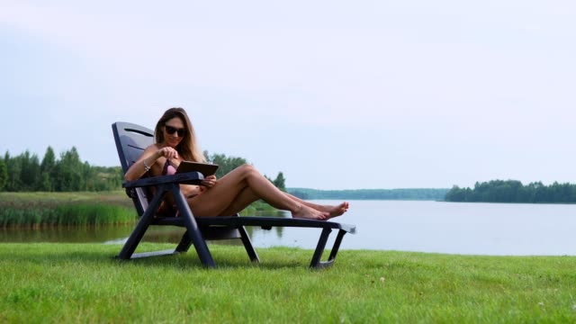 Woman-working-on-a-tablet-computer-traveling-the-world-on-vacation,-sunbathing-on-the-beach-near-the-lake