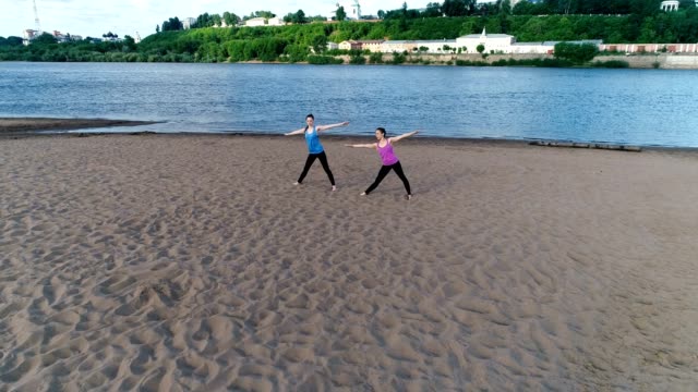 Two-woman-doing-yoga-on-the-sand-beach-by-the-river-in-the-city.-Beautiful-city-view-in-sunrise.