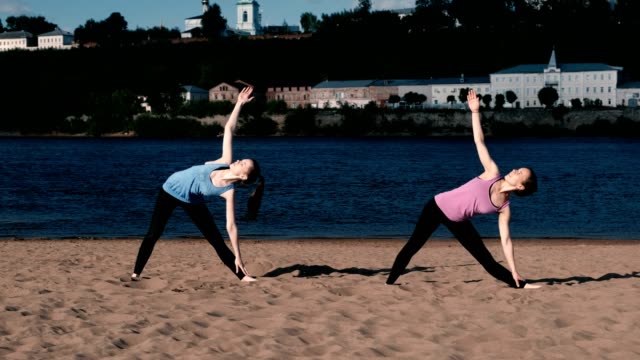 Zwei-Frau-Yoga-am-Sandstrand-am-Fluss-in-der-Stadt-zu-tun.-Blick-auf-die-schöne-Stadt-im-Sonnenaufgang.-Trikonasans-Utiha-darstellen.