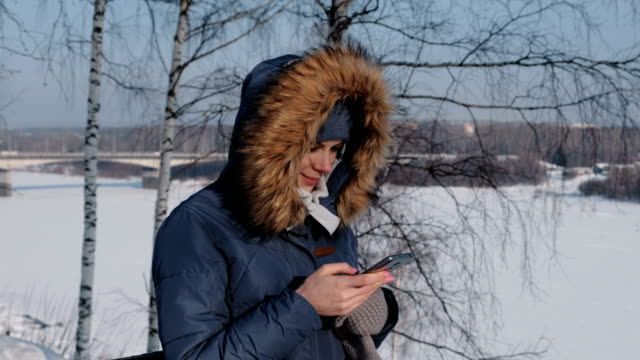 Frau-in-blau-Daunenjacke-mit-Fell-Kapuze-schreibt-messaging-in-ihr-Handy-in-einem-Winter-Park.