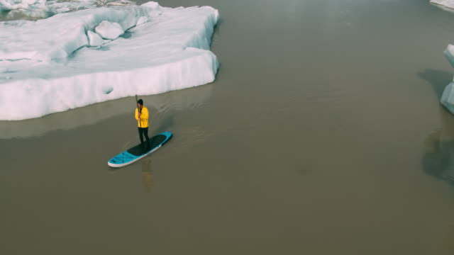 Abenteuerliche-Jüngling-Paddeln-Stand-up-Paddle-Board-durch-Eisberge-im-Gletschersee-in-Island