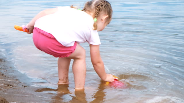 Kleine-Mädchen-holt-und-gießt-Wasser-aus-dem-Eimer-in-den-Sand-Closeup.-Kinder-spielen-am-Strand.