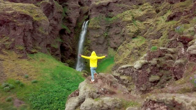 Drone-aerial-view-of-Young-woman-arms-outstretched-at-spectacular-waterfall-in-Iceland--4K-video