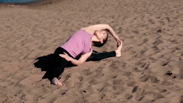 Woman-stretching-yoga-on-the-beach-in-the-city.-Sirsasana,-Supta-upavistha-konasana-pose.