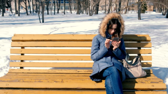 Woman-is-browsing-internet-on-her-phone-sitting-on-the-bench-in-winter-city-park-in-sunny-day.