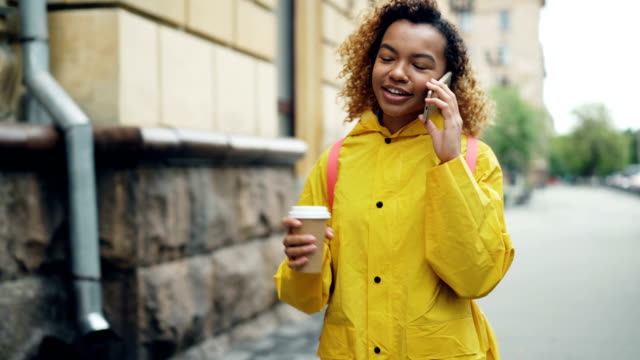 Mujer-bonita-afroestadounidense-es-beber-café-para-llevar-y-charlando-en-el-teléfono-móvil-a-pie-de-la-ciudad-en-primavera-con-chaqueta-brillante.-Concepto-moderno-de-la-tecnología-y-el-Milenio.