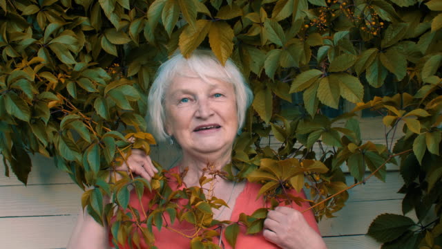 Happy-senior-woman-posing-in-green-leaves-near-wall