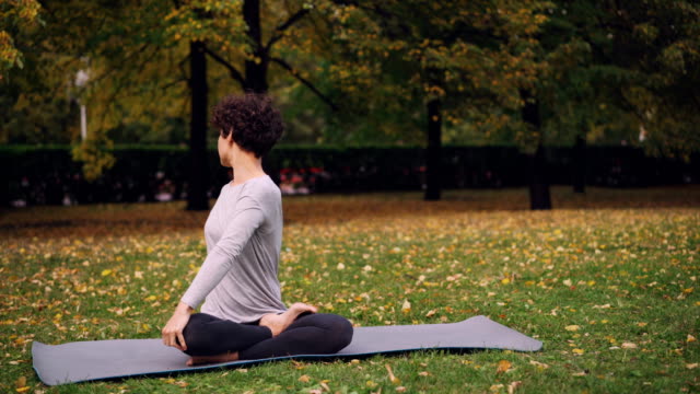 Good-looking-girl-in-sports-clothing-is-twisting-her-body-sitting-in-lotus-position-then-relaxing-with-closed-eyes-after-practice-in-park-on-autumn-day.