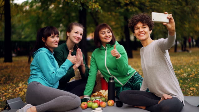 Attraktive-junge-Damen-Yogini-nehmen-Selfie-mit-Smartphone-beim-Picknick-im-Park-im-Herbst.-Mädchen-posiert-und-lächelnd-mit-Spaß-auf-Matten-sitzen.