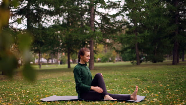 Sonriente-mujer-joven-está-ejerciendo-en-el-parque-haciendo-yoga-sentado-en-la-estera-de-hierba-verde-y-amarilla,-disfrutar-de-aire-fresco-y-saludable-actividad.-Concepto-de-juventud-y-recreación.