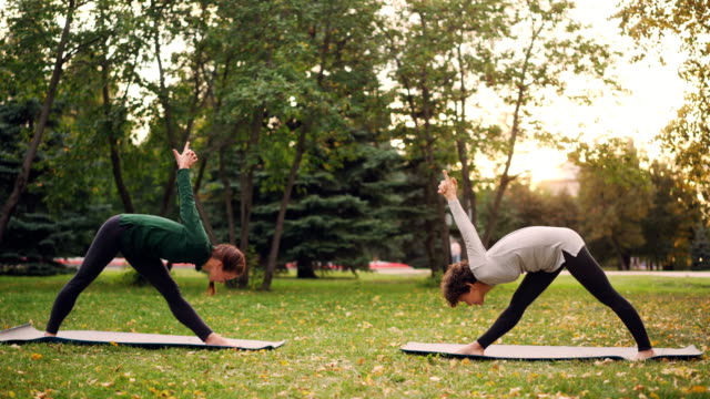 Estudiante-de-yoga-es-hacer-ejercicio-al-aire-libre-con-instructor-de-estiramientos-de-piernas-y-espalda-flexión-hacia-delante-de-pie-sobre-tapetes-en-el-prado-verde-y-amarillo-en-el-parque-de-la-ciudad.