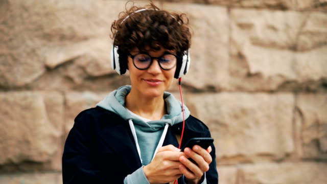 Pretty-girl-is-headphones-and-glasses-is-listening-to-music-and-touching-smartphone-screen-choosing-songs-standing-outdoors-in-city-with-stone-wall-in-background.