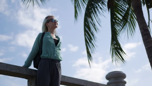 Young-happy-caucasian-woman-with-long-blonde-hair-in-sunglasses-and-green-shirt-standing-and-smiling-near-palm-tree-on-a-blue-sky-background.-Travel-concept