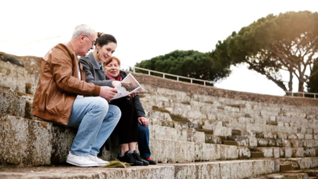Happy-Caucasian-senior-family-with-young-daughter-sitting-and-talking-on-amphitheater-ruins-in-Ostia,-Italy-with-a-map.