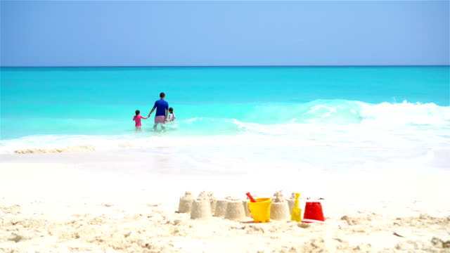 Castillos-de-arena-en-playa-blanca-con-juguetes-de-plástico-de-los-niños-y-la-familia-en-el-fondo-del-mar