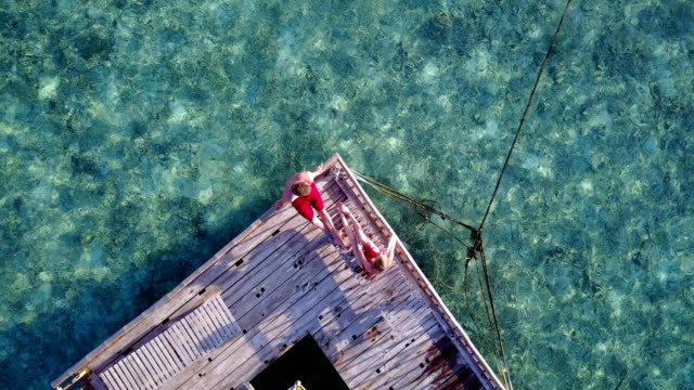 v03825-Aerial-flying-drone-view-of-Maldives-white-sandy-beach-2-people-young-couple-man-woman-relaxing-on-sunny-tropical-paradise-island-with-aqua-blue-sky-sea-water-ocean-4k-floating-pontoon-jetty-sunbathing-together
