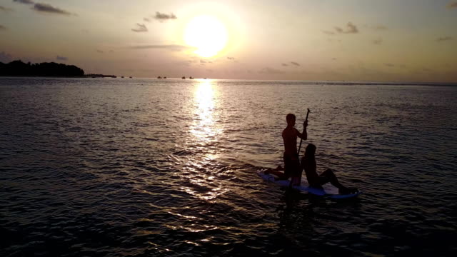 v04076-fliegenden-Drohne-Luftaufnahme-der-Malediven-weißen-Sandstrand-2-Personen-junges-Paar-Mann-Frau-Paddleboard-Rudern-Sonnenuntergang-Sonnenaufgang-auf-sonnigen-tropischen-Inselparadies-mit-Aqua-blau-Himmel-Meer-Wasser-Ozean-4k