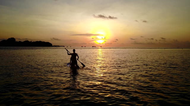 v04094-fliegenden-Drohne-Luftaufnahme-der-Malediven-weißen-Sandstrand-2-Personen-junges-Paar-Mann-Frau-Paddleboard-Rudern-Sonnenuntergang-Sonnenaufgang-auf-sonnigen-tropischen-Inselparadies-mit-Aqua-blau-Himmel-Meer-Wasser-Ozean-4k