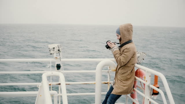 Young-female-journalist-standing-on-the-board-of-the-boat-and-taking-photos-on-the-camera-in-overcast-day