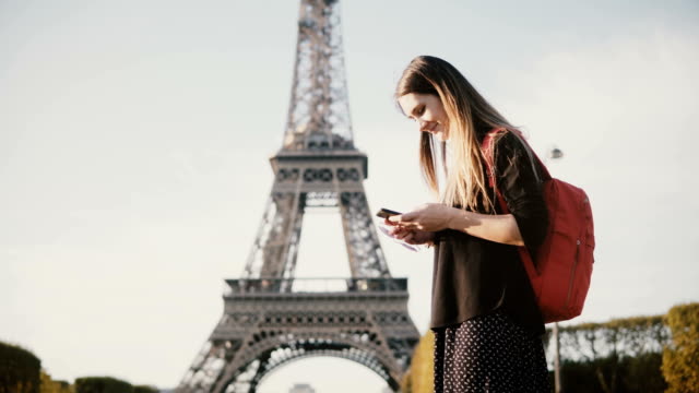 Young-beautiful-woman-with-backpack-standing-near-the-Eiffel-tower-and-using-the-smartphone,-browsing-the-Internet
