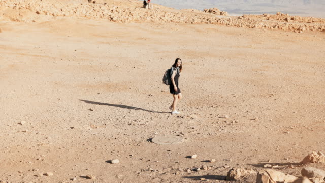 Woman-with-backpack-walks-to-desert-ruins.-Young-Caucasian-female-traveler-on-dry-sand,-rocks,-walls.-Masada-Israel-4K