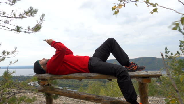 Tourist-man-He-is-lying-on-the-bench-in-the-mountains,-and-using-smartphone-.-men--in-the-woods.-hiking.-a-tour-of-the-forest.-camping.-leisure.-ecotourism.-wild.-adventure-begins
