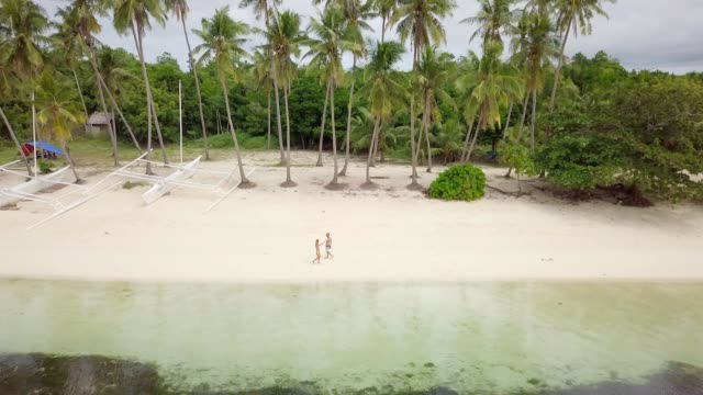 Drone-shot-aerial-view-of-young-couple-walking-on-tropical-beach-in-the-Philippines.-Palm-trees-and-clear-blue-water.-People-travel-love-romance-vacations-concept.-4K-resolution-video