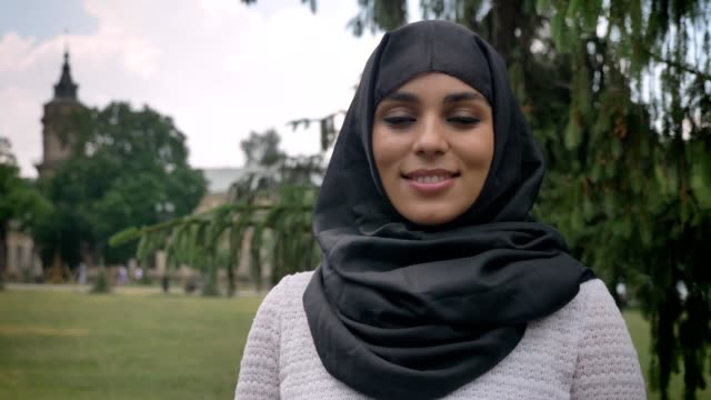 Junge-muslimische-Frau-Hijab-ist-tagsüber-im-Park-bei-schlechtem-Wetter,-lächelnd-gerade-in-die-Kamera,-aufbauend-auf-Hintergrund,-Religiuos-Konzept
