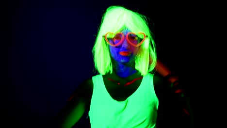 Mujer-con-pintura-UV,-peluca,-gafas-brillantes,-ropa-y-químico-palo-bailando-delante-de-cámara.-Mujer-caucásica.-.