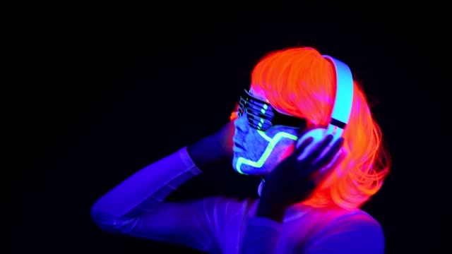 Woman-with-UV-cyborg-face-paint,-wig,-glowing-glasses,-clothing-dancing-and-listening-to-music-with-headphones.-Asian-woman.-.