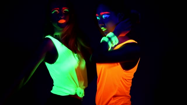 Women-with-UV-face-paint,-glowing-bracelets,-glowing-clothing-dancing-together-in-front-of-camera,-Half-body-shot.-Caucasian-and-asian-woman.-.