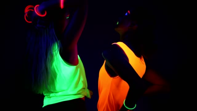 Women-with-UV-face-paint,-glowing-bracelets,-glowing-clothing-dancing-together-in-front-of-camera,-Half-body-shot.-Caucasian-and-asian-woman.-.