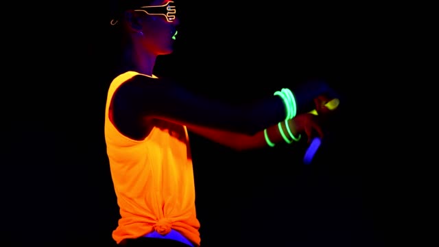 Woman-with-UV-face-paint,-glowing-clothing,-glowing-glasses,-bracelet-dancing-in-front-of-camera-holding-chemical-light,-half-body-side-shot.-Asian-woman.-.