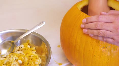 Man-Scoops-Seeds-from-a-Large-Orange-Pumpkin-and-Drops-them-into-a-Metal-Bowl