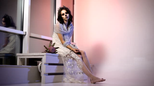 Young-girl-in-white-dress-and-veil-participating-in-halloween-party-sitting-in-the-room.-with-creative-scary-creepy-make-up-is-on-the-bench-near-the-window.-appearance-the-flower