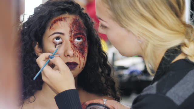 Make-up-artist-que-la-niña-halloween-hacer-upin-estudio.-Arte-de-cara-de-Halloween.-Mujer-se-aplica-a-maquillaje-profesional-en-la-cara-de-chica-española.-Pintura-de-guerra-de-sangre,-cicatrices-y-heridas.-4k