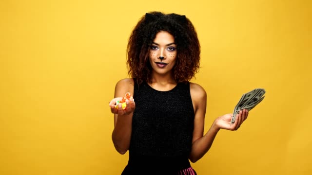 Carefree-african-lady-with-cat-make-up-holding-in-hands-candies-and-money,-trick-and-treat-isolated-over-yellow