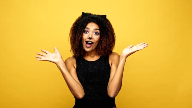 Glücklich-überrascht-Afro-Amerikanerin-mit-Katze-Make-up-und-Ohren-Lächeln-und-Kamera-über-gelbe-Wand-mit-Fledermäusen-isoliert