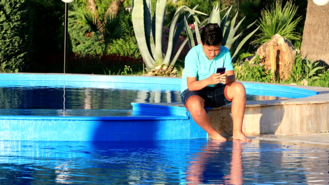 Muchacho-joven-usando-el-teléfono-inteligente-cerca-de-la-piscina