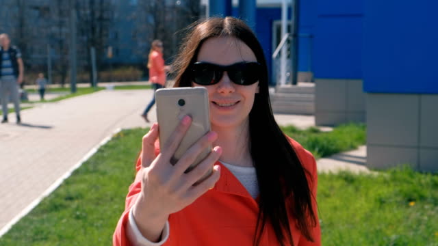Porträt-der-glückliche-junge-Brünette-Frau-in-Sonnenbrille-macht-Selfie-am-Telefon-die-neben-blauen-Gebäude-auf-der-Straße.