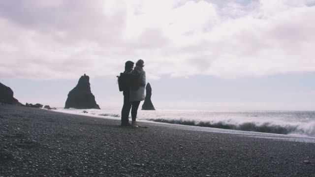 Joven-pareja-cogidos-de-la-mano-en-la-playa-de-arena-negra-en-Islandia,-cámara-lenta