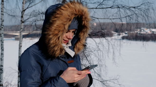 Close-up-woman-in-blue-down-jacket-with-fur-hood-writes-messaging-in-her-cellphone-in-a-winter-Park