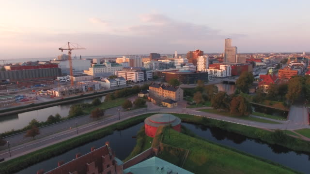 Aerial-view-flying-over-Malmo-city-at-sunset.-Drone-shot-of-"Malmohus"-in-Sweden,-construction-site-and-cityscape-in-the-background