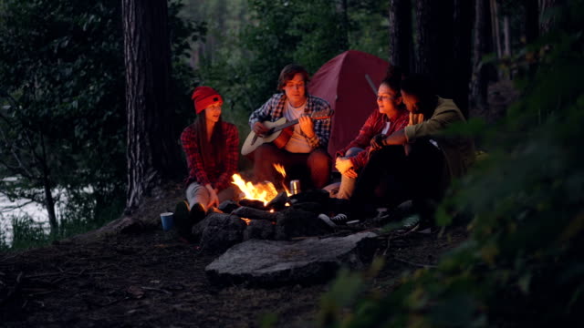 Girls-and-guys-friends-are-singing-songs-while-guitarist-is-playing-musical-instrument-sitting-near-fire-in-forest-and-enjoying-nature-and-music.-Tent-and-lake-are-visible.